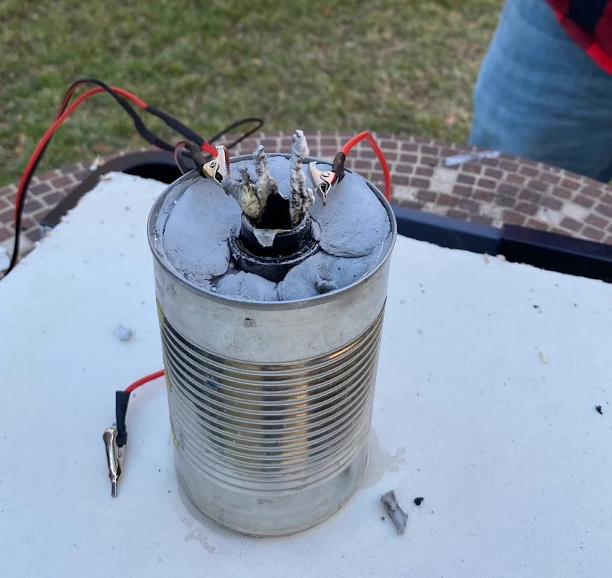 A steel soup can packed with gray clay holding a rocket engine upright, its nozzle at the center, with red and black igniter wires clipped on, standing on a white drywall base.