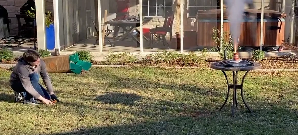 A test firing in a yard: the engine sits on a small round table at the right with smoke rising, while I crouch on the grass at the left.