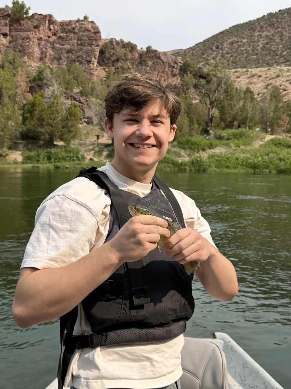 Thomas Kloke smiling on a boat, wearing a life vest and holding a small trout, with red rock cliffs and green hills behind him.
